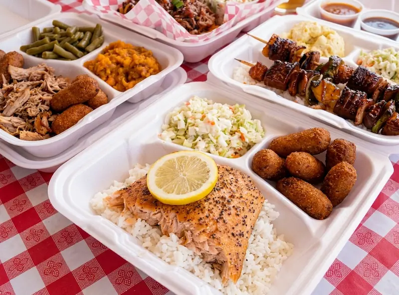 A professional photograph of Southern BBQ food from Smokey and the Pig, featuring a compartment container with baked salmon on rice, plus plates of grilled chicken kabobs and authentic Carolina sides on a red checkered tablecloth.